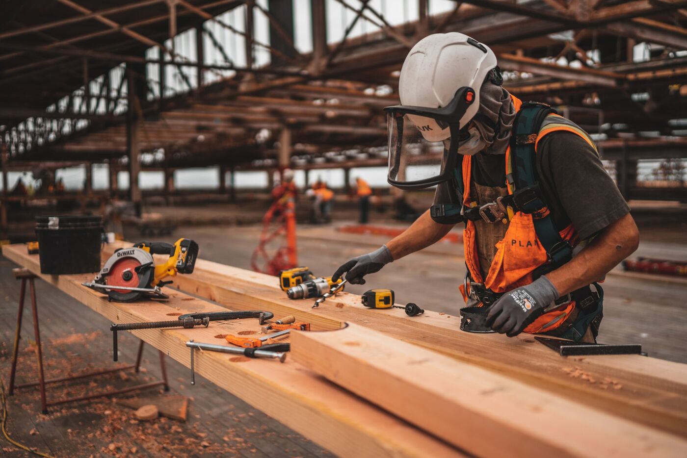 A worker in a face shield and gloves measures timber at a workbench in a factory